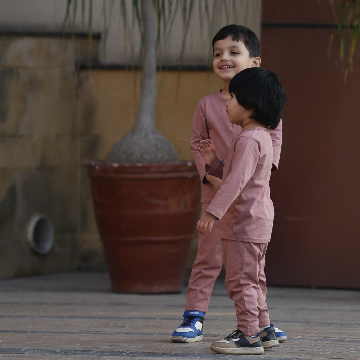Two children in matching outfits standing outdoors with potted plants in the background